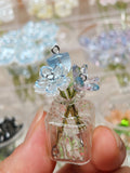 A miniature glass vase charm filled with small blue and light blue flower beads, with a silver jump ring at the top