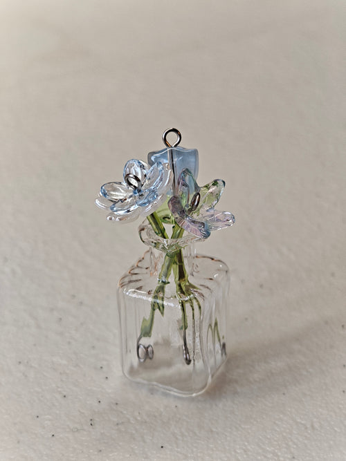 A miniature clear glass vase pendant with two small glass flowers on green stems, photographed on a light beige textured background.